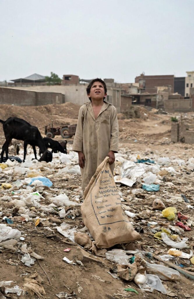 A child stands amid piles of waste in Karachi, holding a sack as he searches for recyclable materials.